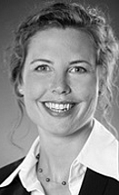 Laura Keders Porträt einer lächelnden Frau mit lockigem Haar, die eine Bluse und einen Blazer trägt. Sie schaut direkt in die Kamera und strahlt Freundlichkeit und Professionalität aus. Der Hintergrund ist unscharf und in Grau gehalten.